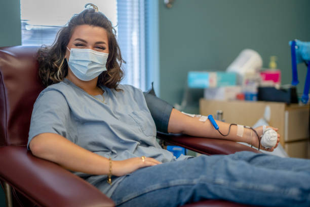 Masked young woman happy to be donating blood A masked young woman is at a hospital for a blood donation drive. She is dressed casually and is seated comfortably on a chair. A medical syringe is hooked onto her arm for the blood withdrawal process. She appears to be happy and smiling behind her mask. blood test stock pictures, royalty-free photos & images