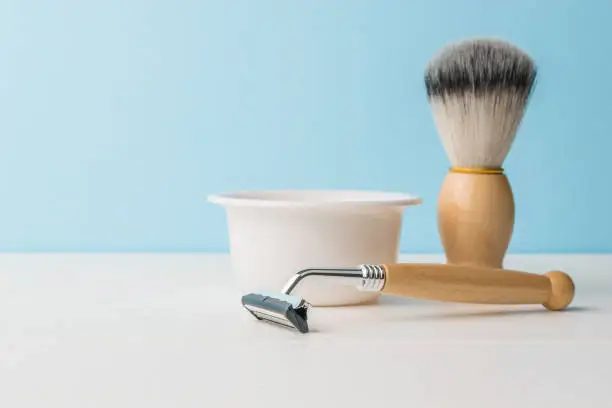 Photo of A ceramic bowl, a shaving brush and a razor with a wooden handle on a white table. Space for the text.