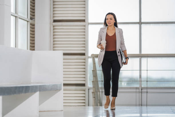 A happy successful Asian businesswoman holds a takeaway coffee cup, document file and tablet walks in the business building stock photo