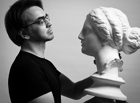 Black and white portrait of adult man sculptor, artist in black t-shirt and glasses holding in hands, looking at antique sculpture woman head over light gray background
