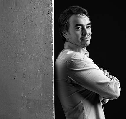 Black and white portrait side view of smirking middle-aged man in shirt, standing back to white concrete wall with arms crossed at chest and looking at camera over black background