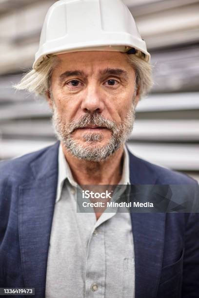 Portrait Of Mature Male Factory Director Wearing White Helmet While Standing Inside Workplace Stock Photo - Download Image Now