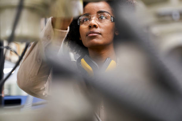 Young female factory operator wearing safety glasses while using machinery control panel inside factory