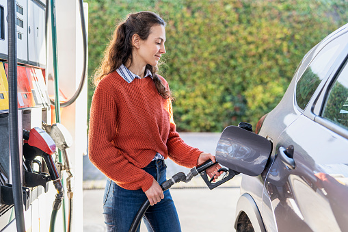 Smiling young adult woman filling fuel tank while standing next to car in filling station during daytime