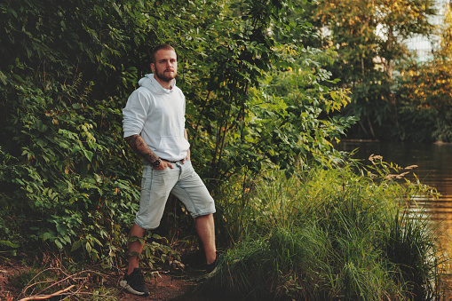 Portrait of young bearded man with tattoos in white jacket in countryside or in park. Male in casual clothes spends evening in nature. Concept of style, walking in fresh air and unity with nature