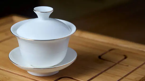 Gaiwan on chaban, selective focus. Tea ceremony gong fu cha. Chinese white pottery Dehua white Chinese porcelain gaiwan bowl tea cup and lid plate with gold border on bamboo teaboard tea tray chaban