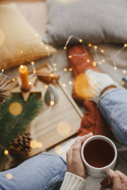Woman hands with warm cup of tea relaxing on soft bed with golden lights, christmas stars, pine, candle and pillows in scandinavian room. Atmospheric moment. Cozy winter holidays at home. Top view manos de mujer con taza de té caliente relajante en la cama suave con luces doradas, estrellas de navidad, pino, vela y almohadas en la habitación escandinava. momento atmosférico. acogedoras vacaciones de invierno en casa. vista superior