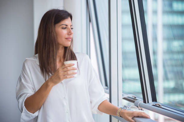 Businesswoman standing by the office window and enjoying her coffee on a break Beautiful businesswoman standing by the office window on a break, having coffee and looking outside 3381 stock pictures, royalty-free photos & images