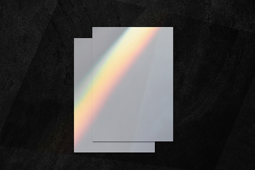 Two empty white vertical rectangle a4 poster, business card or flyer mockups lying on top of each other with overlay of rainbow light refraction caustic effect and shadow on black concrete background.