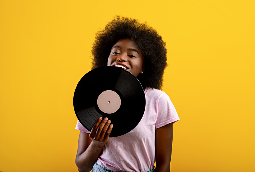 Joyful african american woman holding vinyl record, smiling and looking at camera, standing over yellow background, copy space. Retro music concept.