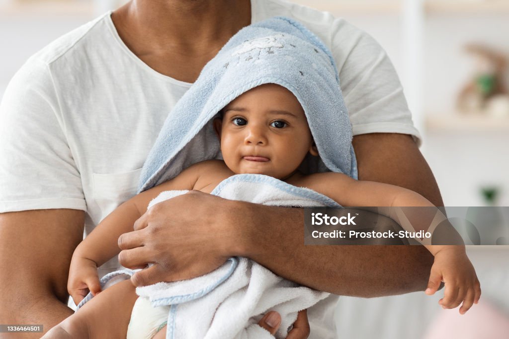 Adorable Black Infant Baby In Towel Relaxing In Father's Arms After Bath Adorable Little Black Infant Baby Wrapped In Blue Towel Relaxing In Father's Arms After Bath, Cute African American Toddler Child Looking At Camera, Enjoying Daddy's Care, Cropped Shot, Closeup Baby - Human Age Stock Photo