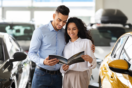 Loving arab couple checking booklet in car salon, choosing new family automobile, handsome middle-eastern man and curly woman standing among luxury autos in showroom, holding brochure