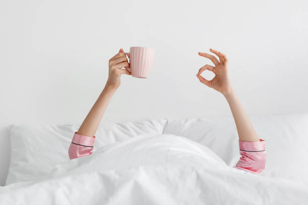 Weekend or vacation, good morning with delicious coffee Weekend or vacation, good morning with delicious coffee. Woman showing arms raised up, holding cup under duvet in bedroom, lady with two hands sticking out from blanket, wake up with fun show ok sign protruding stock pictures, royalty-free photos & images