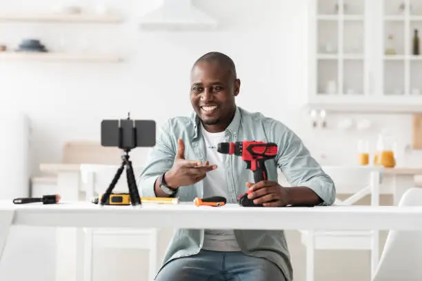 Photo of African american handyman recording video review of instruments, showing electric screwdriver and talking to smartphone