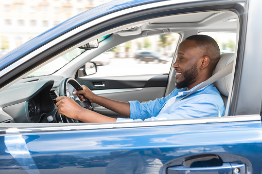 Happy Driver. Side view profile portrait of cheerful positive African American man sitting in a car on driver's seat. Excited black guy riding in the city, holding hands on steering wheel