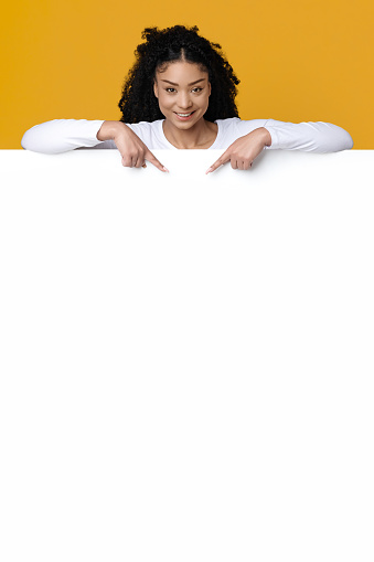Place For Ad. Cheerful Young Black Lady Pointing Down At Big White Advertisement Board, Smiling African American Woman Demonstrating Free Copy Space On Blank Billboard, Standing On Yellow Background