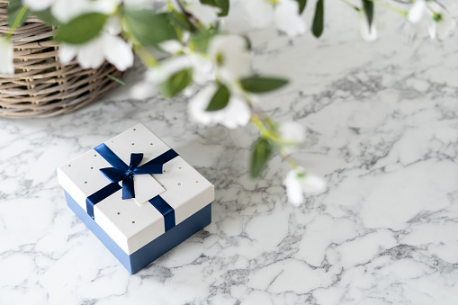 Gift box with ribbon and bow under luscious flowers, white and blue colours, big woven basket with greenery, marble table surface, empty space for text, blurred foreground