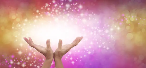 female cupped hands reaching up into a beautiful white light against a gold and pink energy field background with sparkles and white light