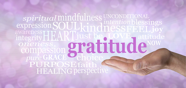 Male hand with the word GRATITUDE floating above surrounded by a relevant word cloud against a pink purple bokeh background