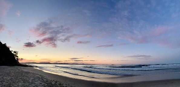 Horizontal landscape panorama of Wategos Beach with waves breaking onto the sand and the sunset clouds and sky reflecting on the wet sand.