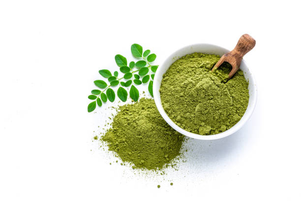 Matcha tea powder and leaves on white background Overhead view of Matcha tea powder in a white bowl isolated on white background. Some Matcha powder is spilled on the background and Moringa leaves are beside the bowl. A wooden serving scoop complete the composition. High resolution 42Mp studio digital capture taken with Sony A7rII and Sony FE 90mm f2.8 macro G OSS lens moringa leaves stock pictures, royalty-free photos & images