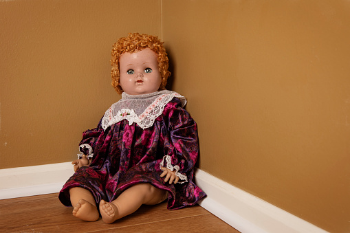 A creepy vintage baby doll sitting in a corner on the floor.