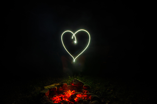 Man drawing a heart with light of mobile phone  in front of campfire during summer night