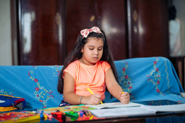 linda niña estudiando en casa, haciendo dibujo o proyecto de arte con colores y crayones mientras está sentada en el sofá. - schoolgirl little girls crayon human face fotografías e imágenes de stock