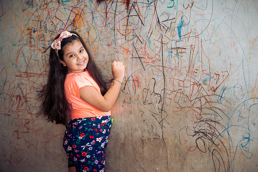 Indoor image of Asian/ Indian Funny cute little girl drawing with marker or crayons on wall at home. Young artist painting on wall at living room