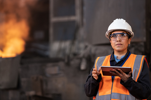 Front view of woman wearing safety equipment and watching logistic processes in big steel factory. Production factory, metal industry, engineering and people at work concept.