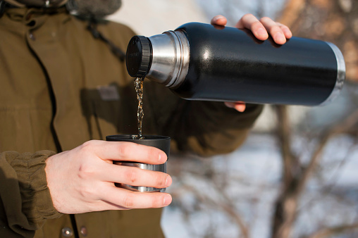 male tourist pours hot tea from a thermos in the winter on the street. Winter hiking concept, camping, travel. close-up.