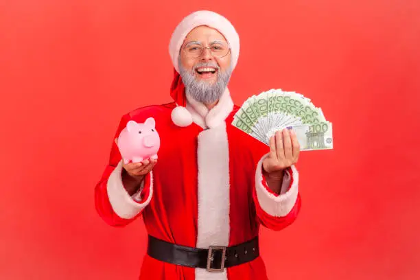 Photo of Positive elderly man with gray beard wearing santa claus costume standing holding euro banknotes and piggy bank, looking at camera with smile.