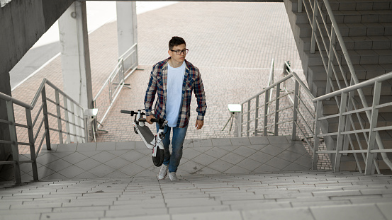 A student climbs the stairs to the university with an electric scooter. modern city concept. Ecological technological lifestyle. Man carries an electric scooter in a folded position
