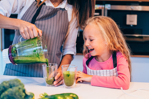 Mother with daughter making fresh smoothie in the kitchen. Beautiful young woman and cute blond girl using blender for green healthy smoothie. Mother pouring green veggie juice in glass for her daughter.