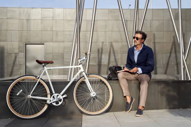 Adult businessman sitting in a notebook writing in a notebook. He uses a bicycle to get around the city. Green transportation concept. High quality photo