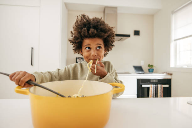 Parental Influence on Eating Behavior Cute child with afro eating spaghetti out of a big yellow pot in the kitchen. Child eating food with his hands. kids eating noodles stock pictures, royalty-free photos & images