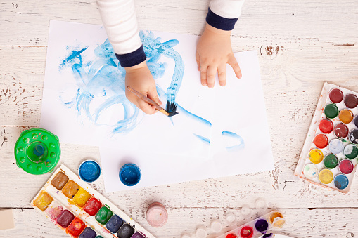 A creative space for drawing and embodying creative ideas. Early learning. Education concept. Top view of a child's hands drawing on paper. Watercolors, brushes and artistic things on a white table.