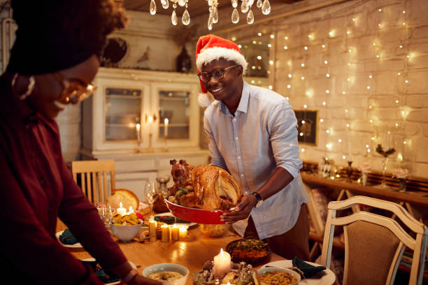 istock Happy black man bringing roast turkey while setting dining table with is wife on Christmas. 1336546304