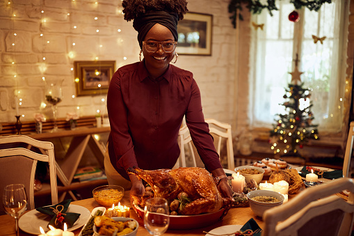 Young happy black woman setting the table for Thanksgiving lunch and serving roast turkey while looking at camera.