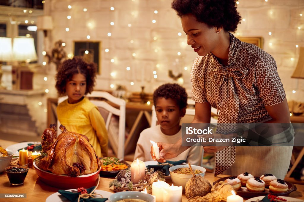 African American mother lights candles during Thanksgiving meal at dining table. Single black mother celebrating Thanksgiving with her children and lights candles during dinner at dining table. Thanksgiving - Holiday Stock Photo