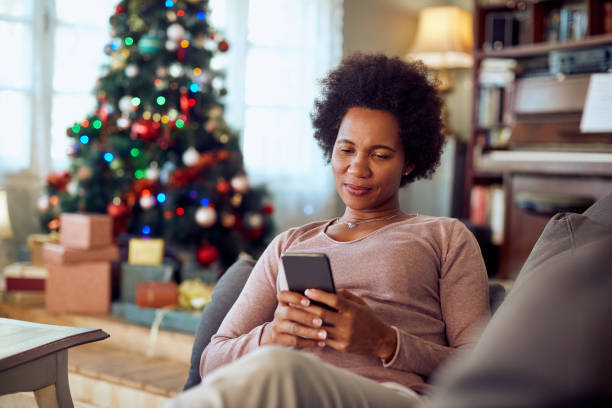 istock Happy black woman text messaging on mobile phone while relaxing on the sofa during Christmas day. 1336546466