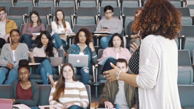 A mid adult female college professor gestures as she teaches an attentive group of diverse students.