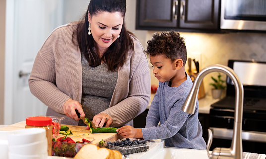 A mother and her son standing in the kitchen preparing food for lunch. The boy is trying to help her slice vegetables on a cutting board. Mom is Caucasian, in her 30s and the boy is mixed race Black and Caucasian, 3 years old.