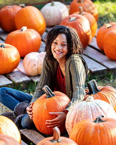 A 12 year old African-American girl sitting on the ground with lots of big pumpkins, smiling at the camera on a sunny autumn day. She is at the pumpkin patch picking out a pumpkin for halloween.