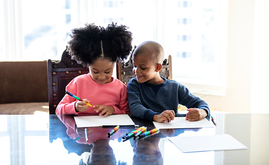 Two African-American children, siblings, at home, sitting at the dining room table, drawing pictures. They are side by side. The 7 year old boy is watching as his 8 year old sister starts to  draw.