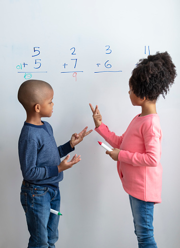 Two African-American children, siblings, standing at a large whiteboard with markers doing math. They are working together to solve addition problems. The 7 year old boy and 8 year old girl are counting on their fingers.