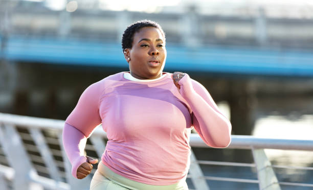 African-American woman with large build, running in city A mid adult African-American woman in her 30s exercising in a city park on the waterfront. She is a plus size model with a large build, wearing long sleeved sports clothing. She is running, jogging or power walking toward the camera with a serious expression. curvy black women stock pictures, royalty-free photos & images