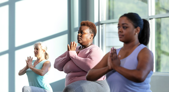 A multi-ethnic group of three women taking a yoga class, in a crescent lunge pose on knee with prayer hands. They are indoors, side by side, with the focus on the African-American woman in the middle. She is in her 40s.