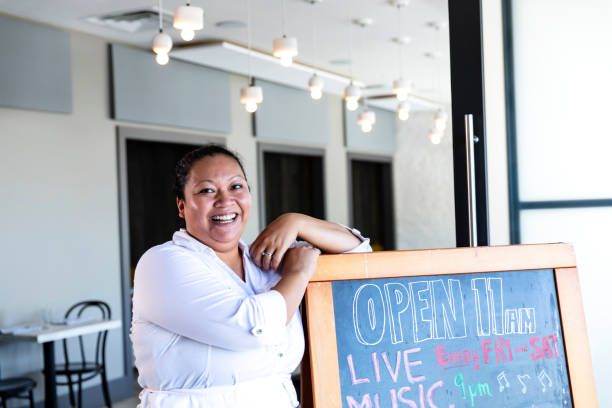 Restaurant manager standing next to open sign A restaurant manager standing beside a sign showing the opening time, smiling at the camera, leaning on the board. She is a mid adult woman in her 30s, mixed race Filipino and Cambodian. cambodian ethnicity stock pictures, royalty-free photos & images