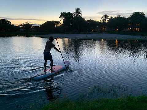 Fit Latin Tourist male doing Paddle boarding at a lake in Miami, Florida, USA, enjoying and relaxing at the sunset time. He is wearing a blue board short.